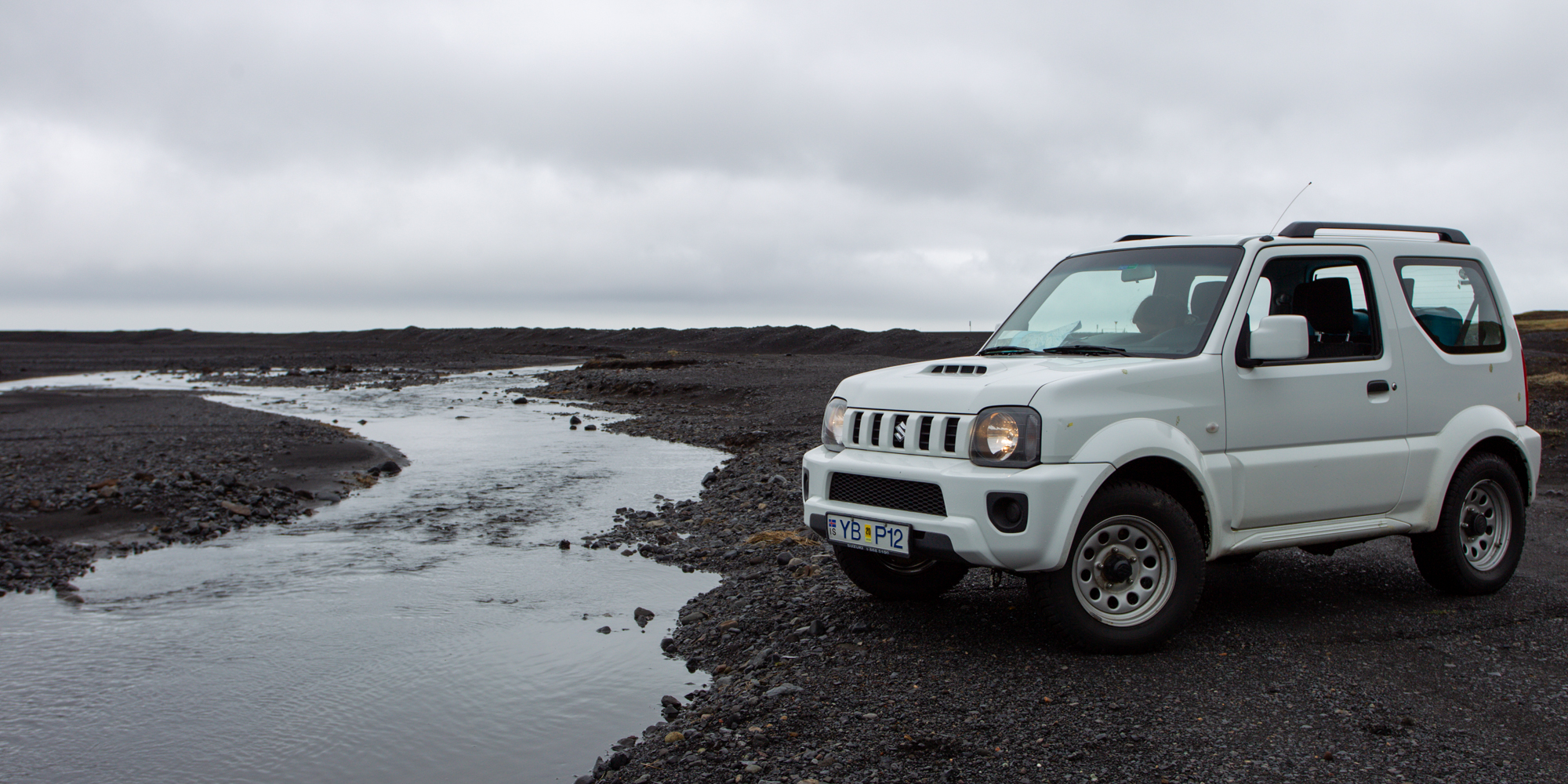 Suzuki Jimny geparkeerd bij een gletsjerrivier op zwart zand in IJsland